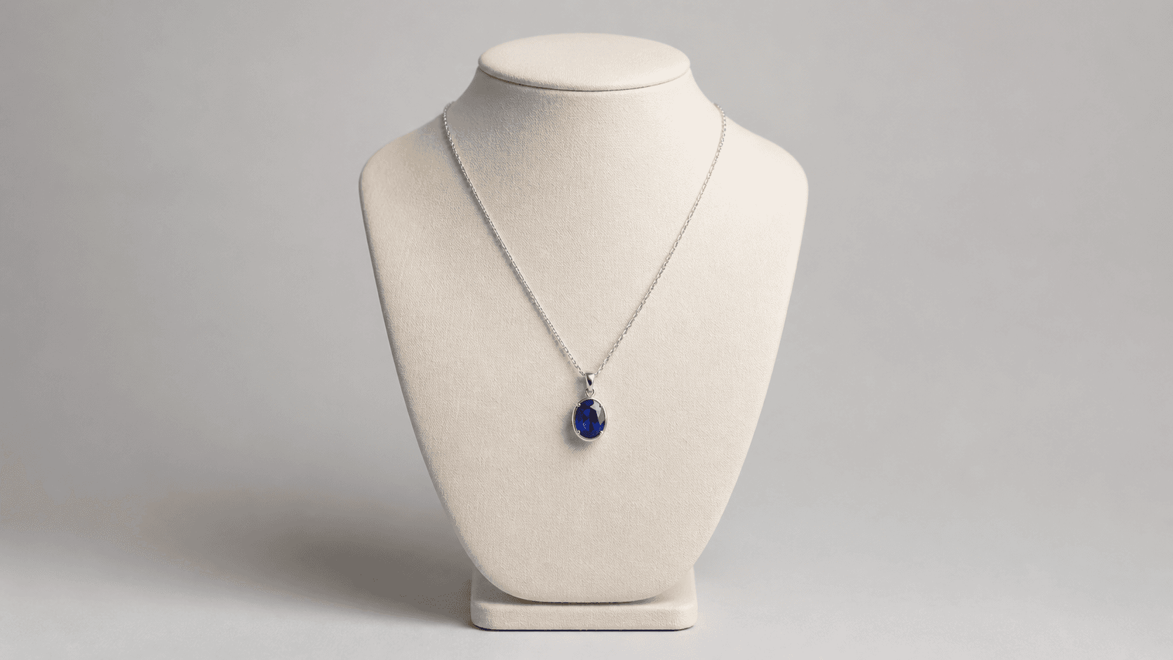 A silver necklace with a blue pendant presented on a cream jewelry display against a neutral studio background